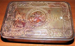A metal tin with carved border and centre cameo; the letters M are on either side.