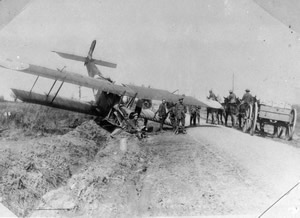 A plane lies nose down in a ditch as soldiers wait nearby and others on horseback and driving a horse and cart pass by.