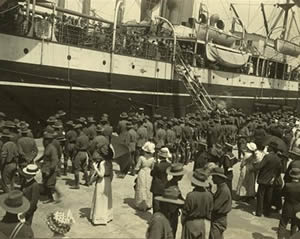 Troops and civilians stand on a wharf in front of a docked ship